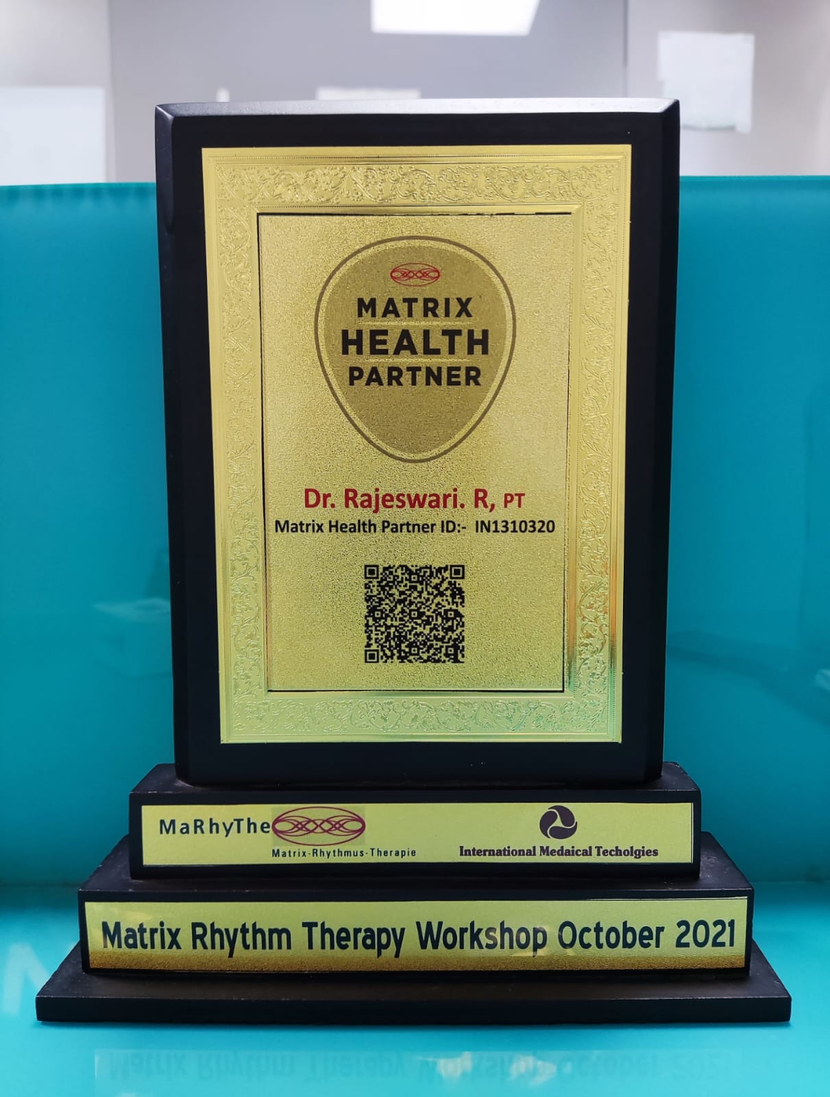 A black framed plaque with golden details and text that reads "MATRIX HEALTH PARTNER" and "Dr. Rajeswari. R, PT" along with a QR code and an ID number, sitting on a stand that states "Matrix Rhythm Therapy Workshop October 2021" and features two logos, one for "MaRhyThe" and another for "International Medical Technologies". The plaque is placed on a turquoise colored table with a white background.